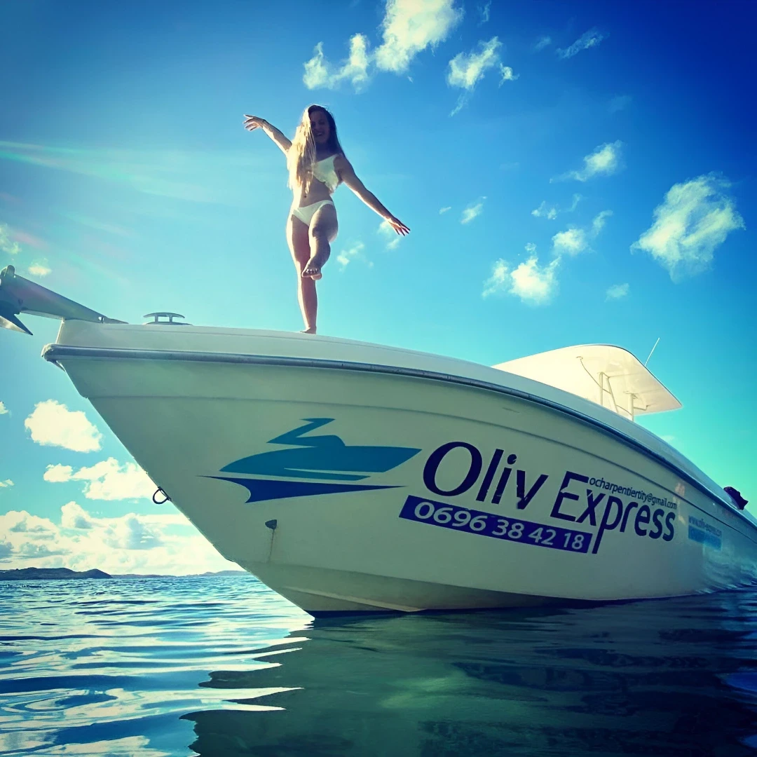 Personne en maillot en équilibre sur la proue d’un bateau Oliv Express, au-dessus d’une eau turquoise sous un ciel ensoleillé