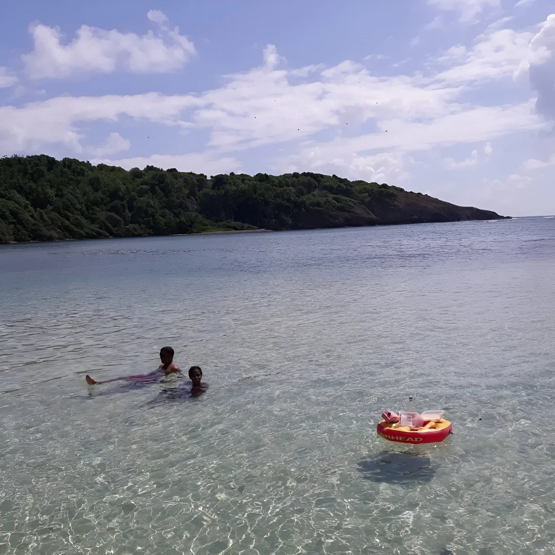 Deux enfants se baignant dans une eau transparente au bord d’une baie tropicale, avec un îlot boisé à l’arrière-plan