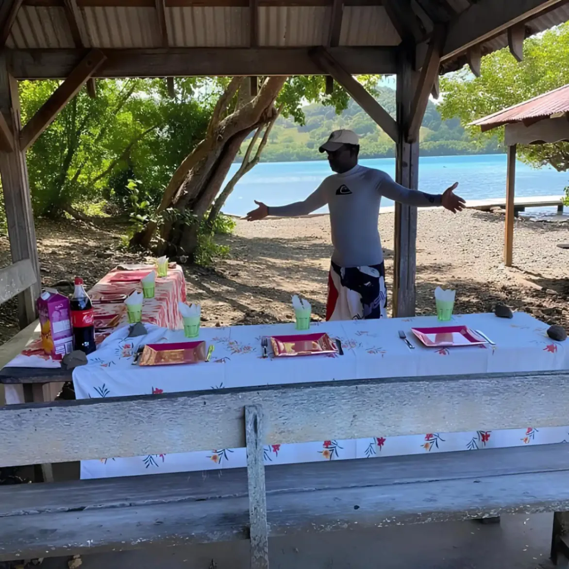 Personne accueillant devant une table dressée pour un pique-nique sous un abri en bois, près de la mer dans un cadre naturel