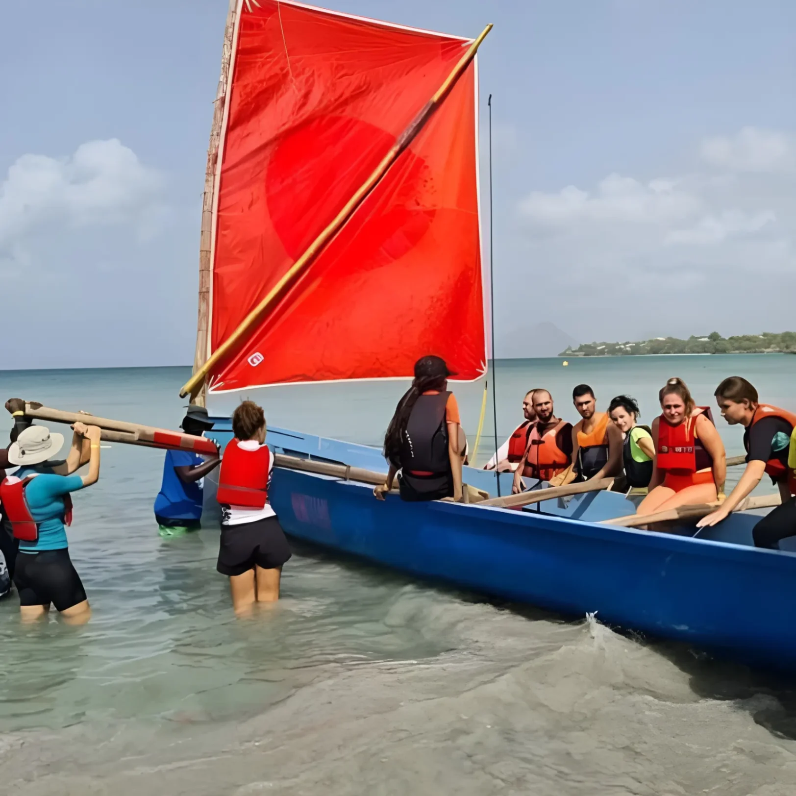 Participants en gilet de sauvetage aidant à mettre à l’eau un bateau traditionnel à voile rouge sur une plage calme