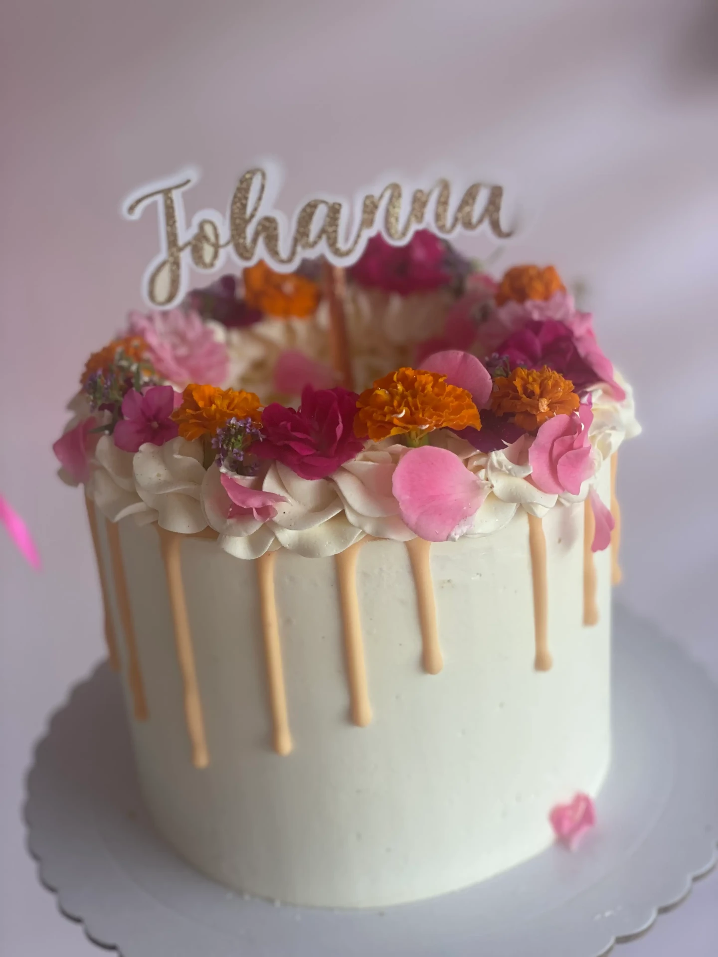 Layer cake blanc décoré de fleurs fraîches colorées et d’un topper “Johanna”, avec effet drip beige