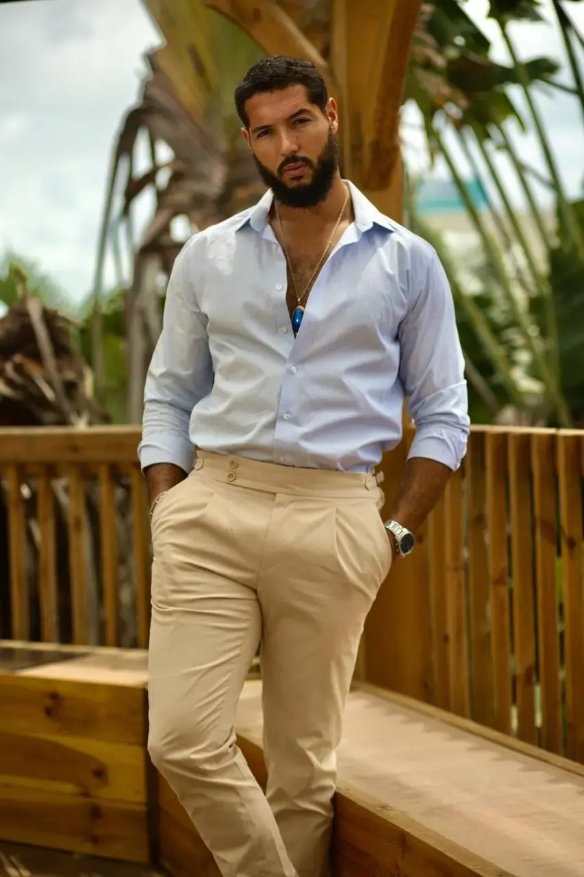 Homme élégant portant une chemise bleu clair et un pantalon beige, posé sur une terrasse en bois entourée de végétation tropicale.