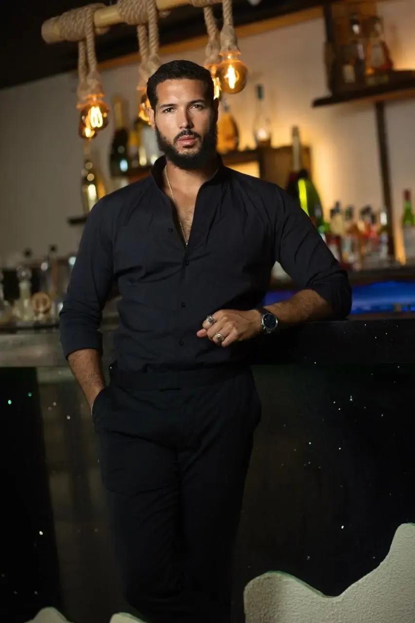 Homme habillé entièrement en noir avec une chemise et un pantalon chic, posé dans un bar éclairé de lumières chaudes.