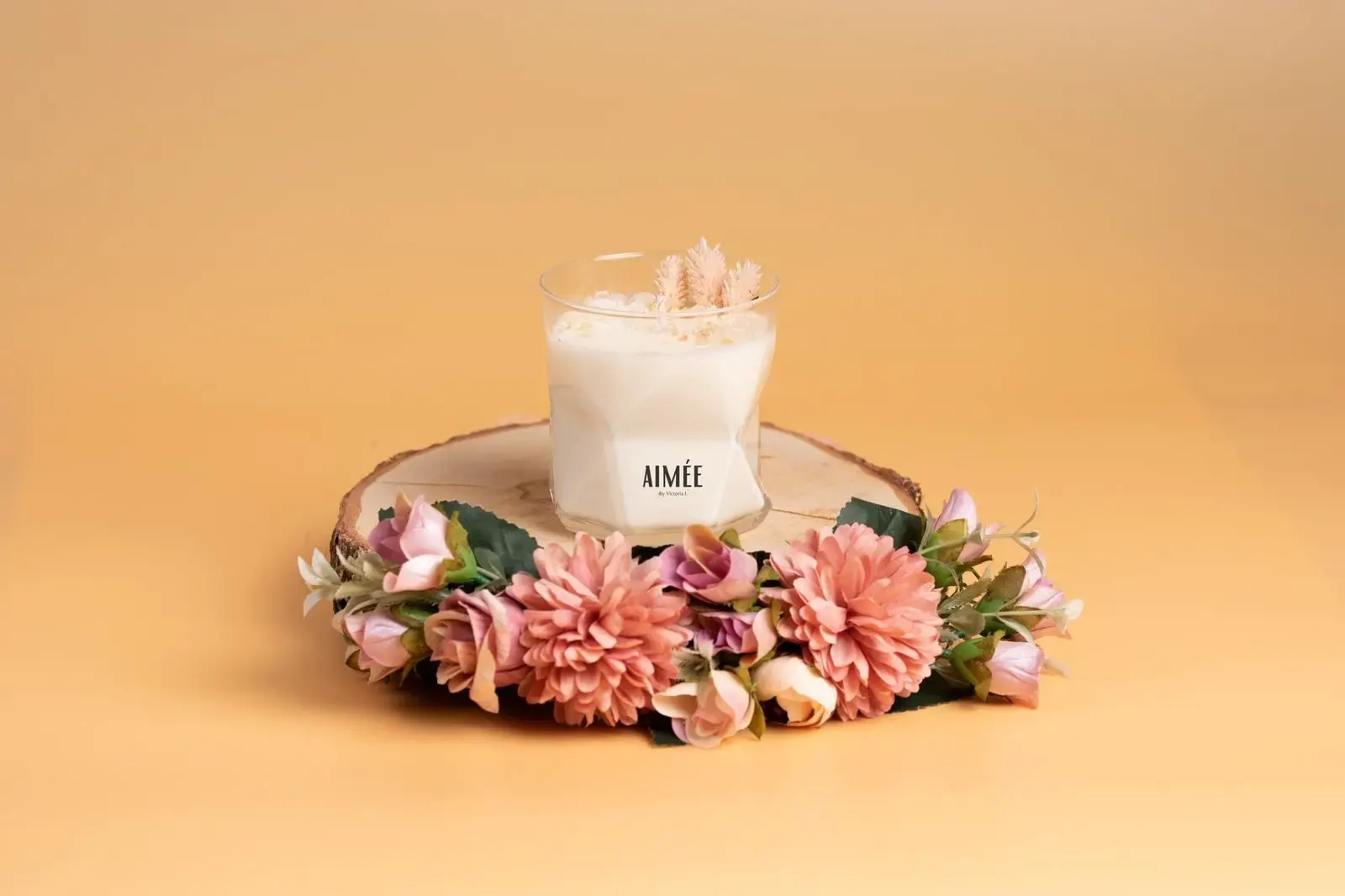 Bougie artisanale AIMÉE dans un verre transparent posée sur un rondin de bois, entourée d’une couronne de fleurs aux tons roses et pêche