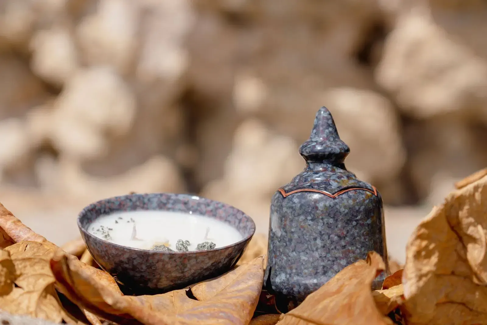 Bougie artisanale blanche dans un bol en céramique mouchetée, posée sur des feuilles sèches avec son couvercle assorti