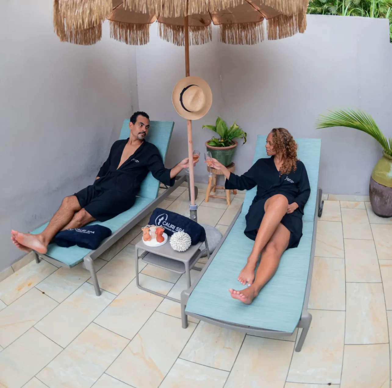 Couple en peignoirs noirs profitant d’un moment de détente sur des transats sous un parasol, trinquant ensemble.