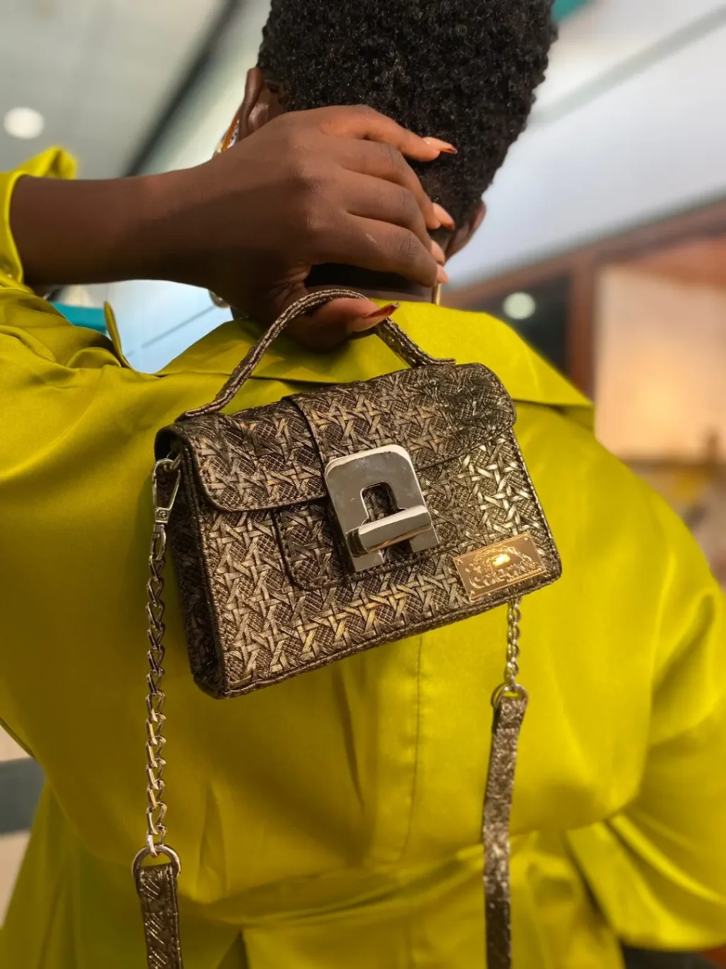 Petit sac doré texturé avec chaîne et grand fermoir métallique, porté sur l’épaule d’une personne en tenue satinée jaune.