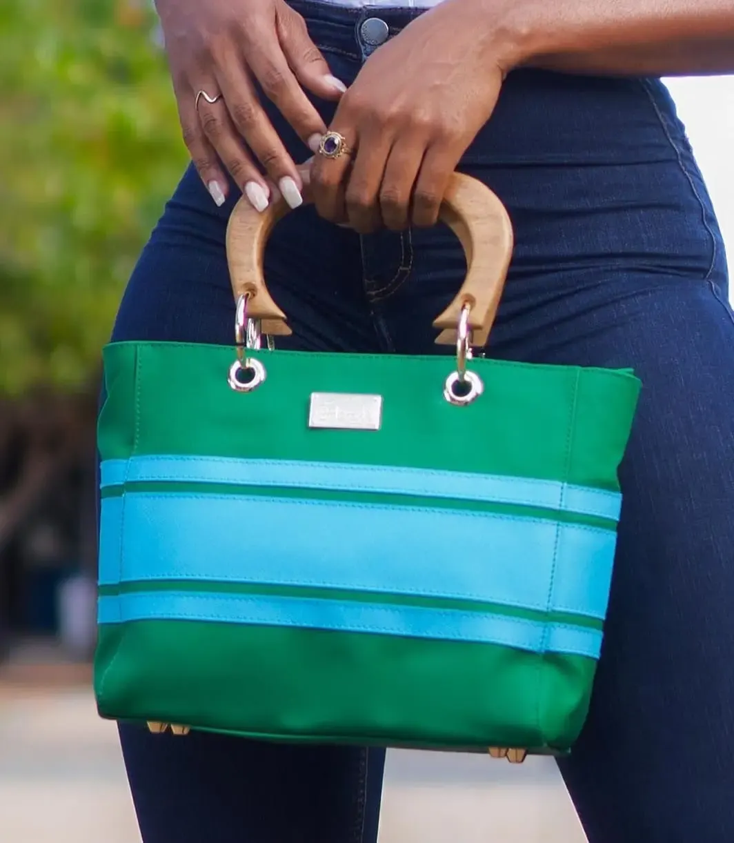 Sac à main vert et bleu avec anses en bois, tenu à la main par une personne portant un jean foncé.