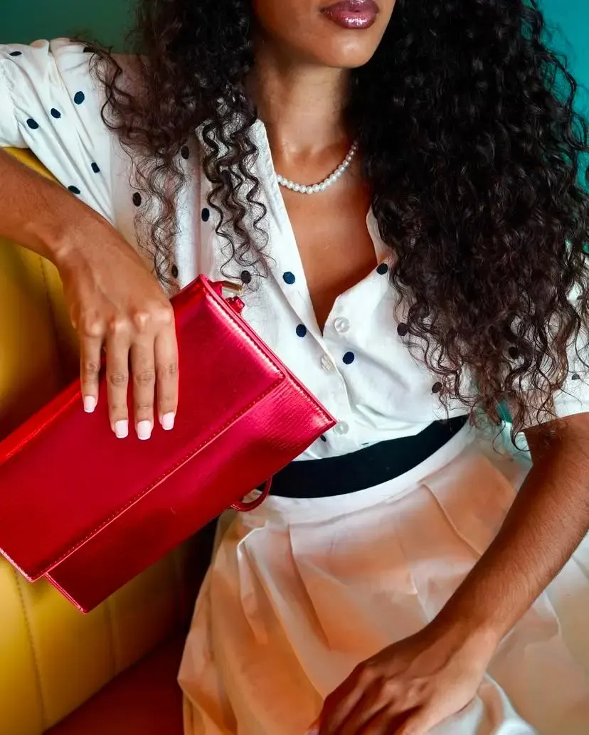 Femme portant une chemise blanche à pois et tenant un sac métallisé rose vif.