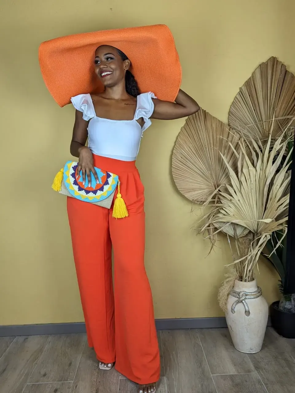 Femme portant un grand chapeau orange avec un top blanc et un pantalon large assorti, look estival élégant et coloré