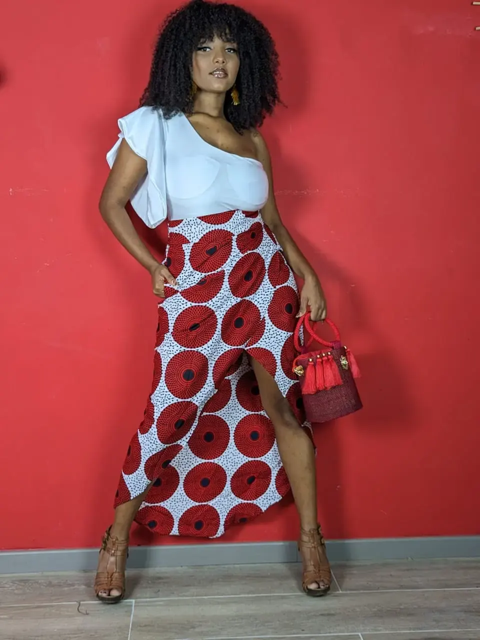 Femme portant une jupe wax rouge et blanche avec un top blanc asymétrique, look élégant et moderne devant un mur rouge