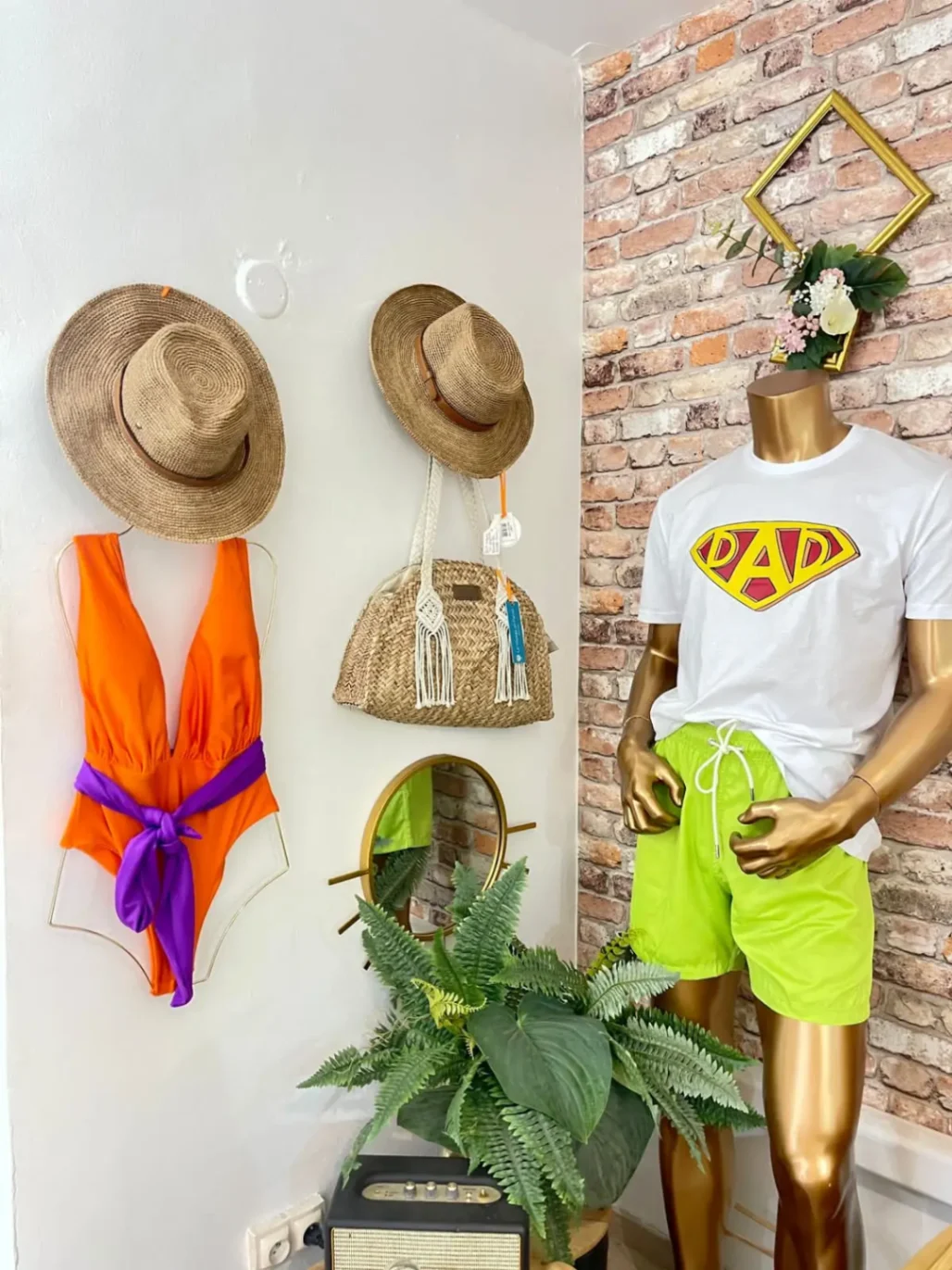 Présentation boutique : mannequin doré portant un t-shirt blanc et short vert, maillot de bain orange exposé avec chapeaux de plage et accessoires estivaux.