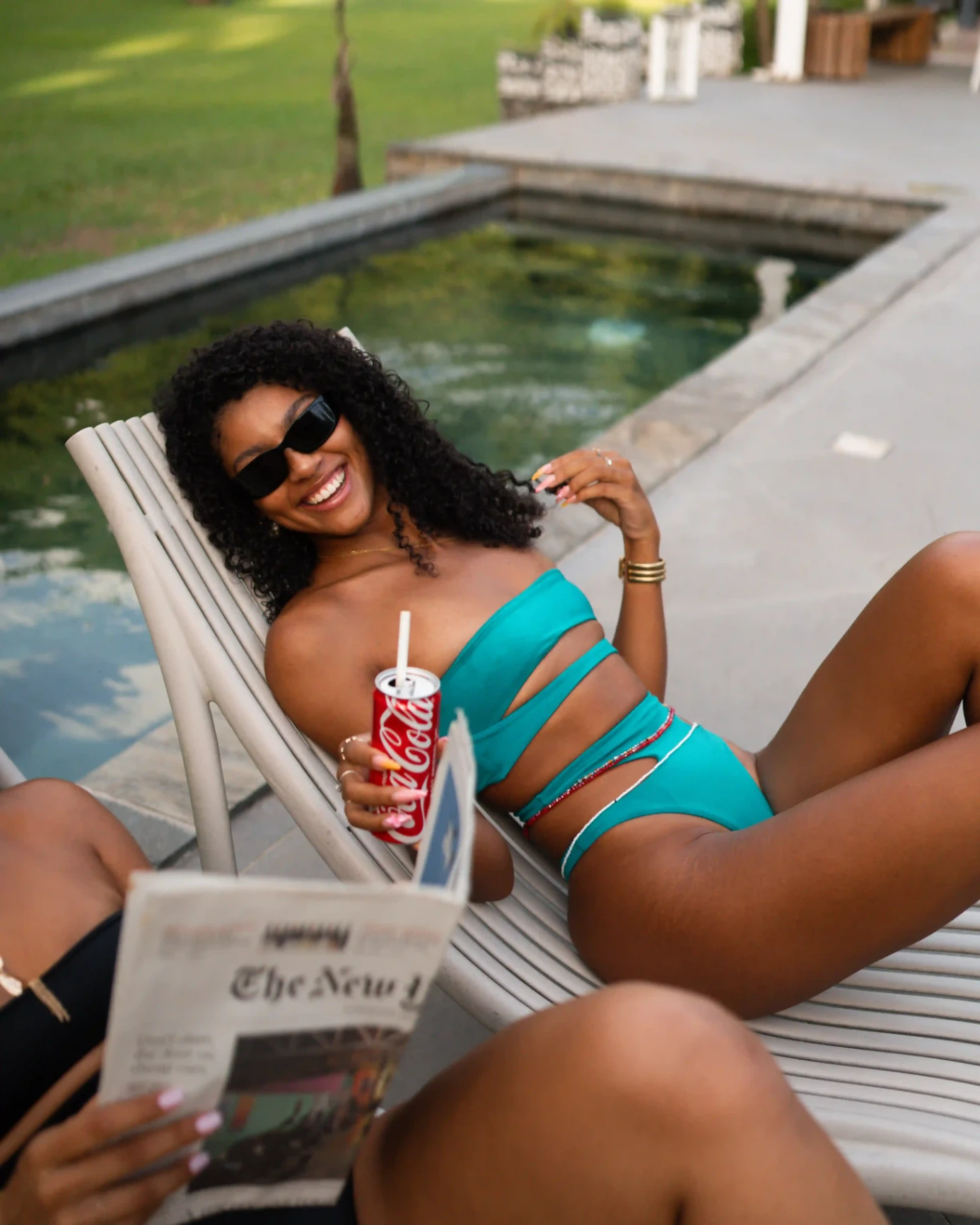 Femme portant un maillot de bain turquoise, allongée sur un transat près d’une piscine et souriant tout en tenant une boisson gazeuse.