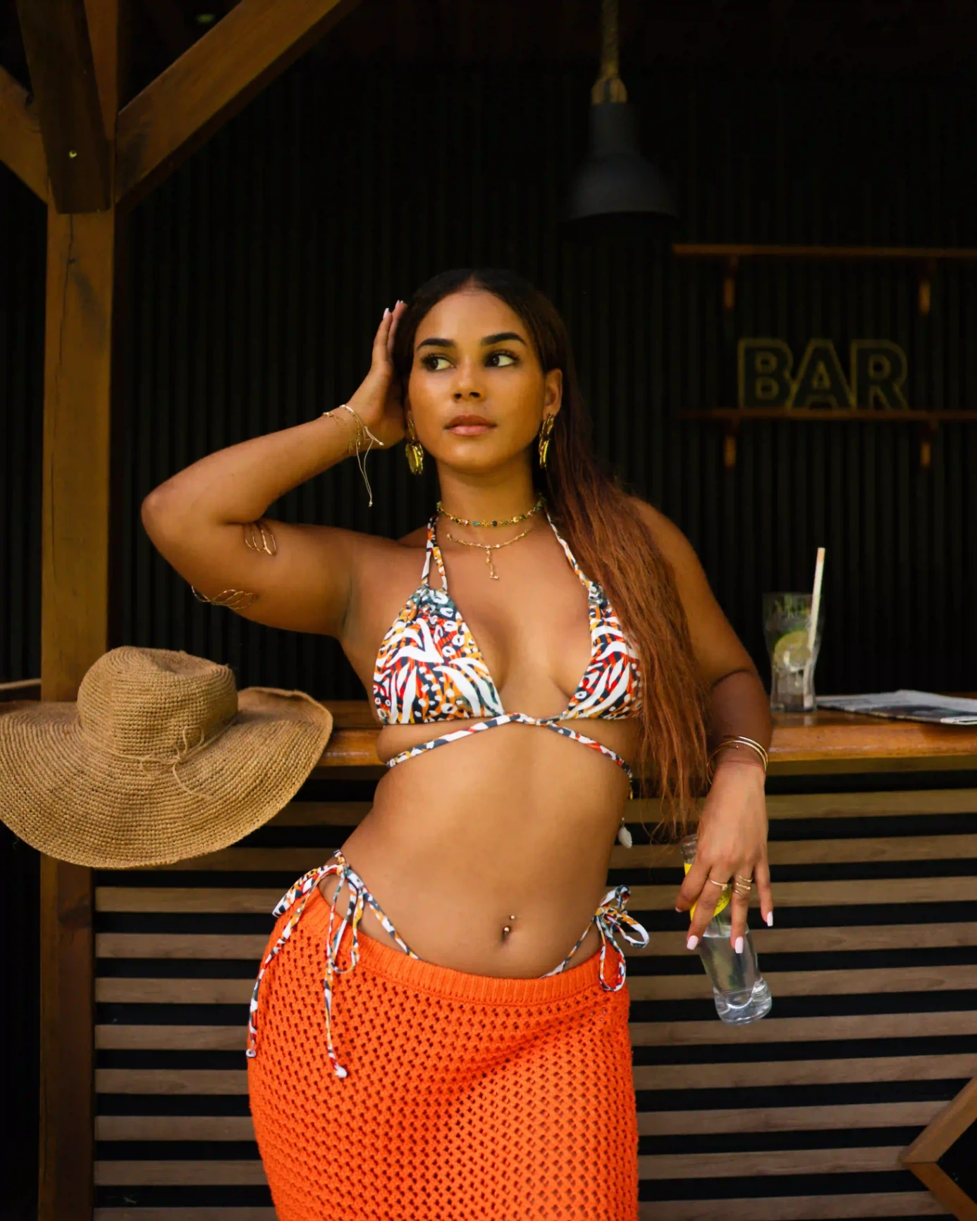 Femme portant un bikini à imprimé coloré, posant devant un bar extérieur en bois avec un chapeau posé à côté.