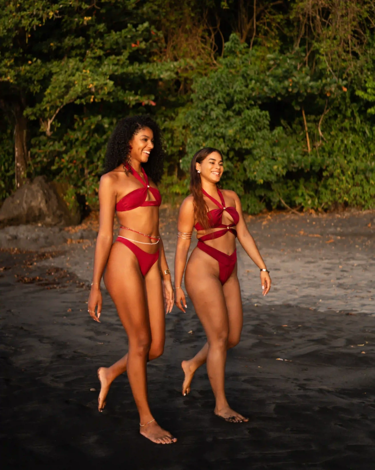Deux femmes marchant sur une plage de sable foncé, portant des maillots de bain rouges assortis dans un décor tropical.