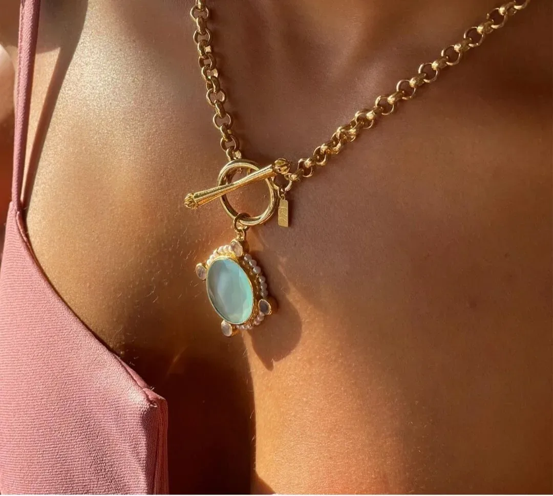 Femme portant un collier doré avec un pendentif bleu nacré, éclairé par la lumière du soleil.