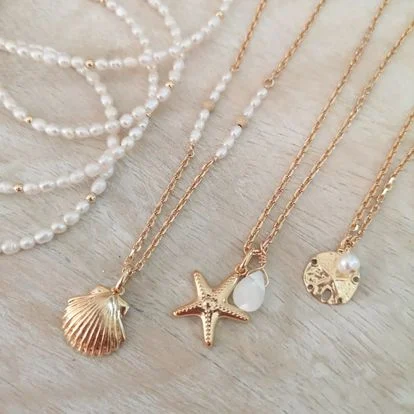 Colliers dorés avec pendentifs coquillage, étoile de mer et médaille perlée posés sur une surface claire.