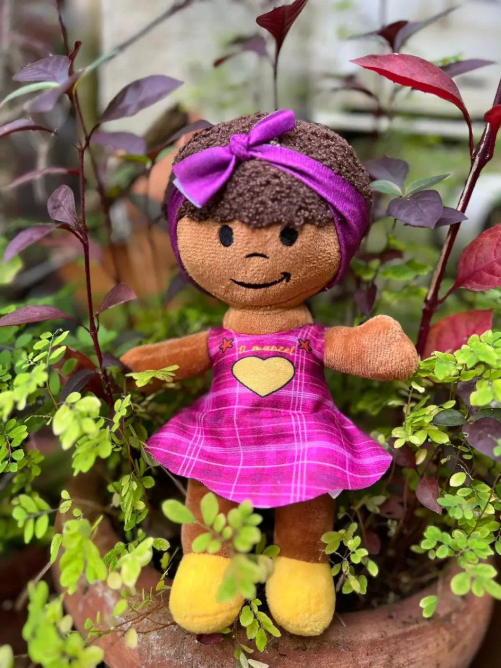 Peluche Ti-Manzel en robe rose et bandeau violet, assise dans un pot entouré de végétation.