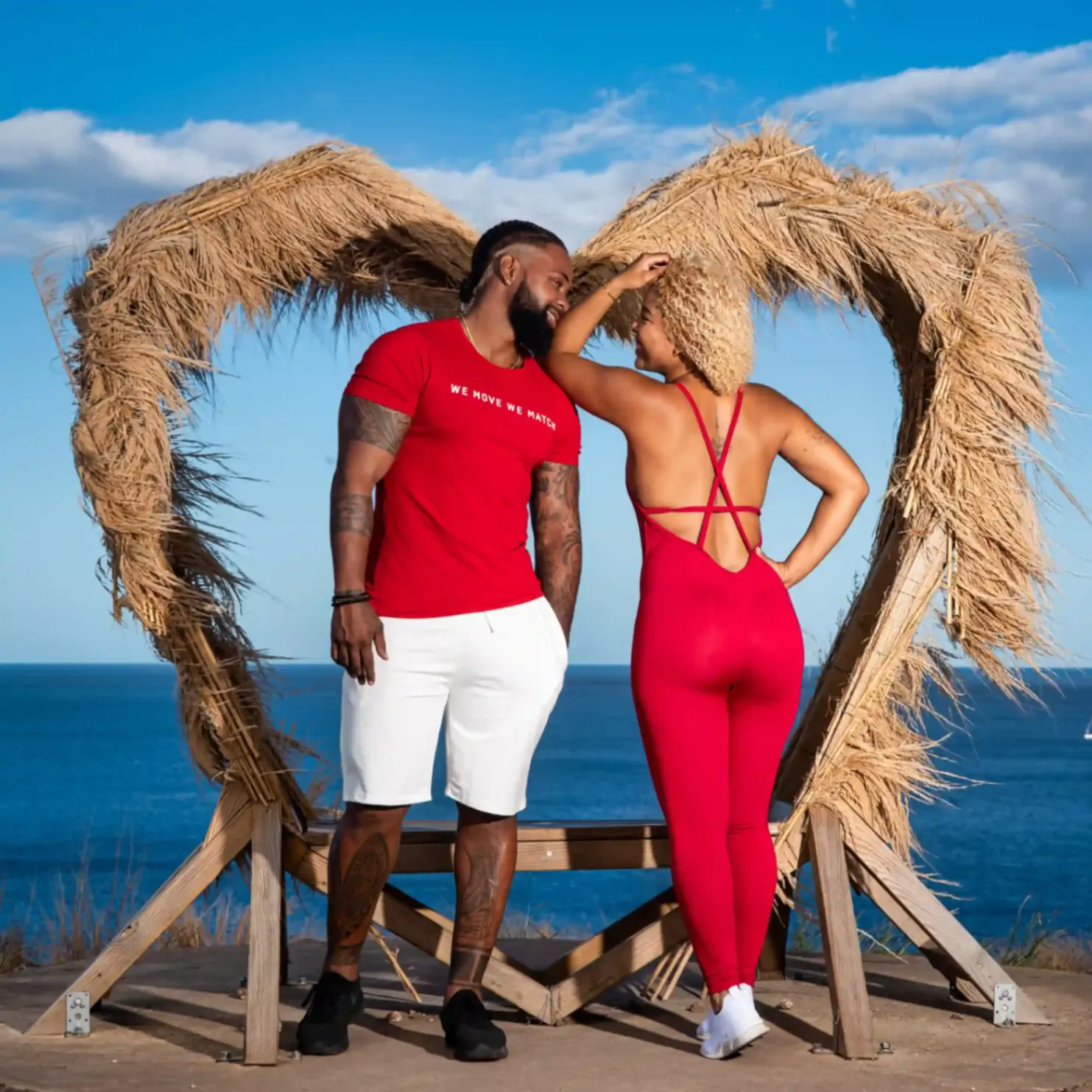 Couple en tenue de sport rouge posant devant une sculpture en forme de cœur au bord de la mer.