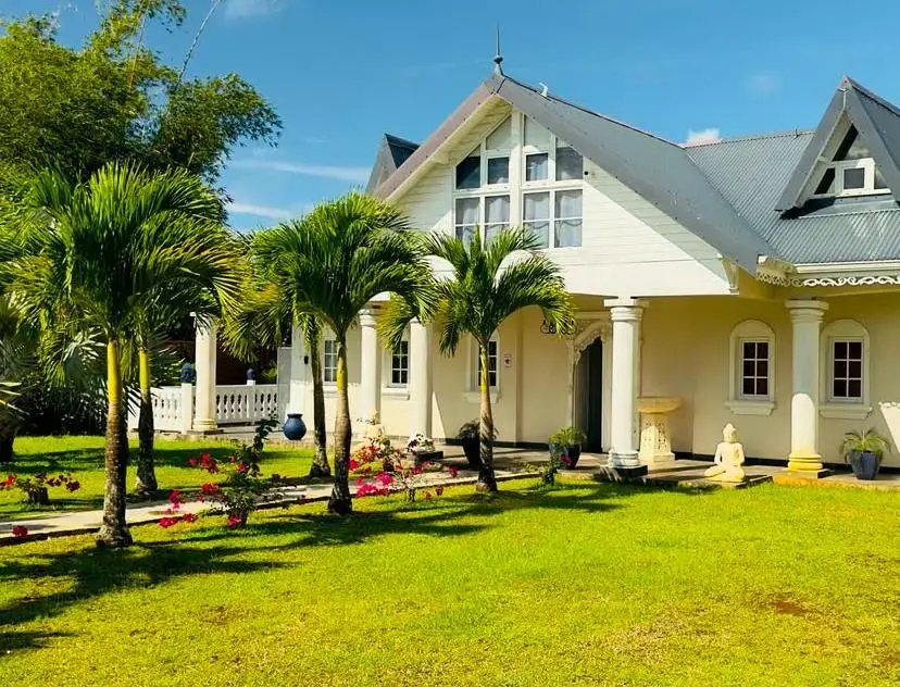Grande villa blanche entourée de palmiers et d’un jardin fleuri sous un ciel bleu.