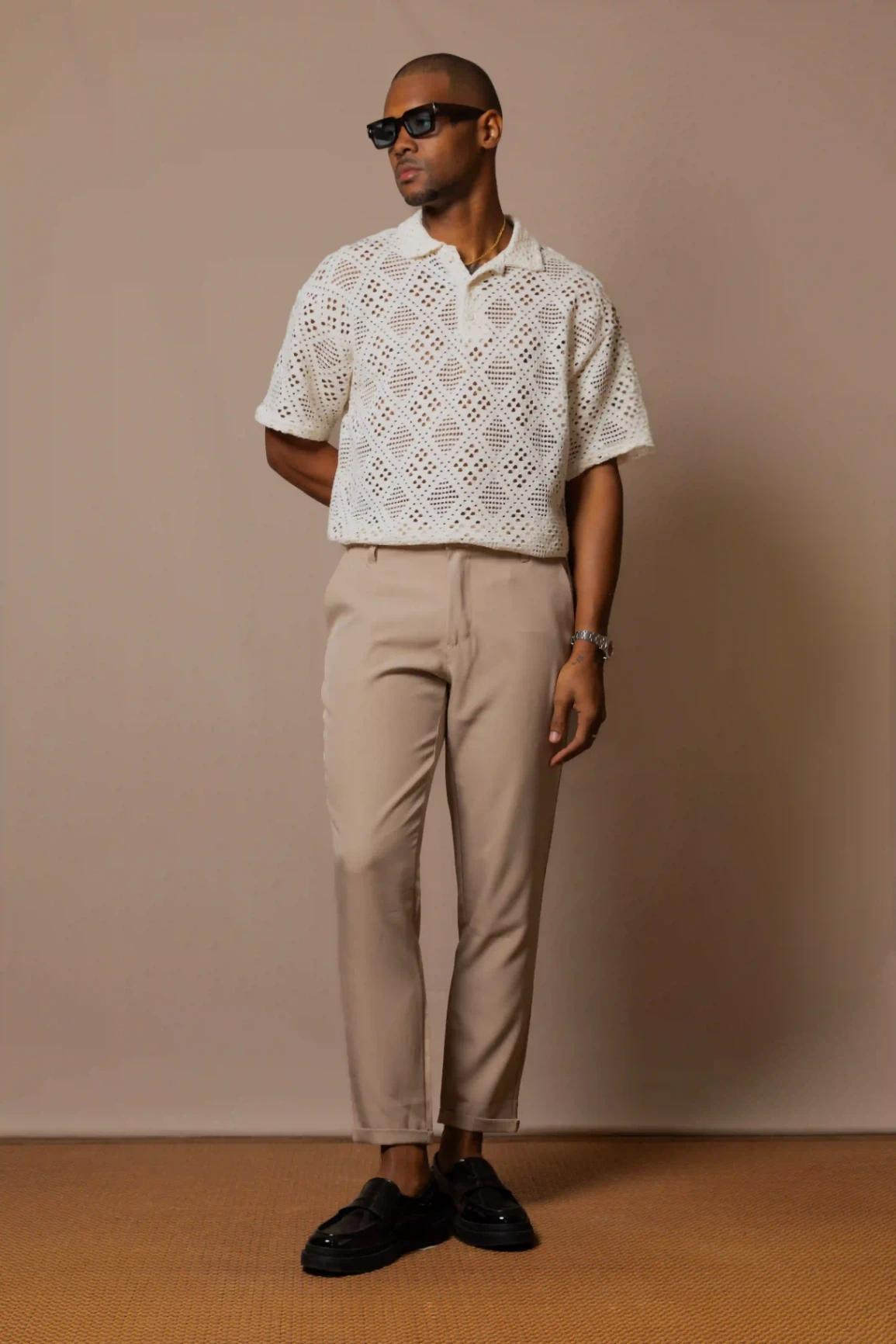 Homme vêtu d’une chemise ajourée blanche et d’un pantalon beige, posé devant un fond neutre.