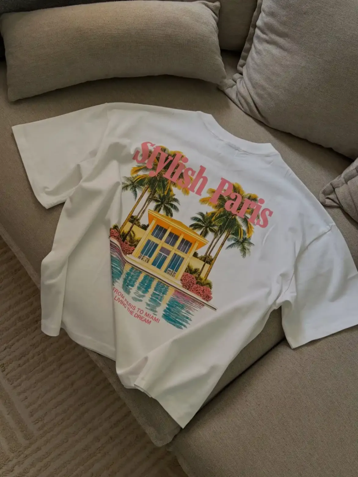 T-shirt blanc Stylish Paris posé sur un canapé avec une illustration de maison tropicale et palmiers au dos.