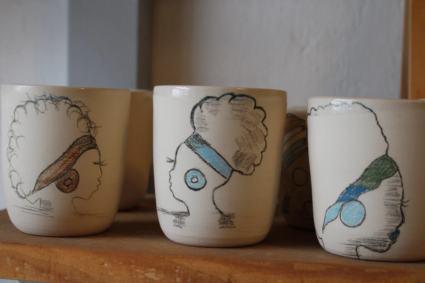 Tasses artisanales en céramique décorées de profils africains stylisés, réalisées à la main et exposées sur une étagère.