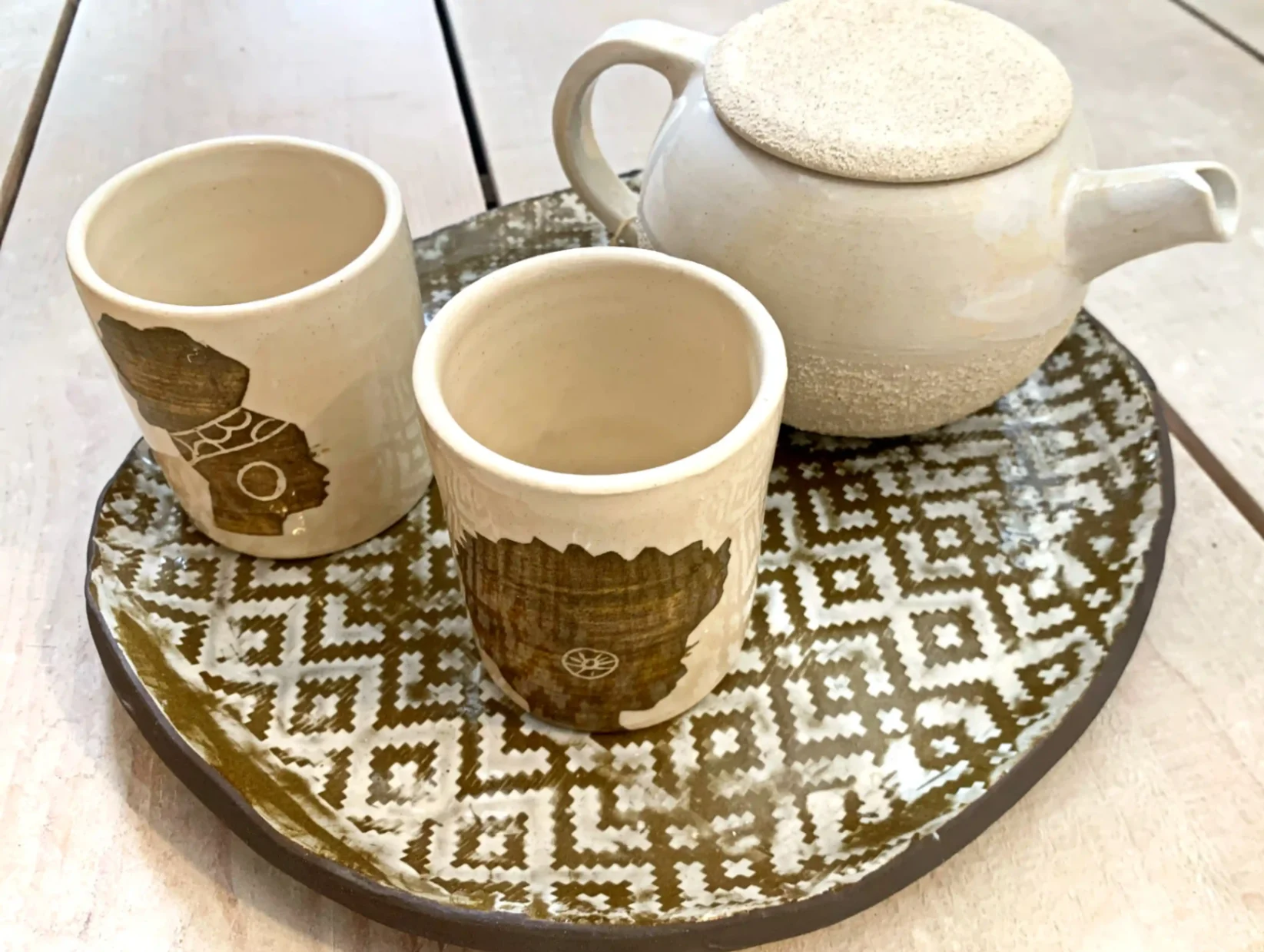 Service à thé artisanal en céramique comprenant une théière et deux tasses décorées de silhouettes africaines, présenté sur un plateau aux motifs géométriques.