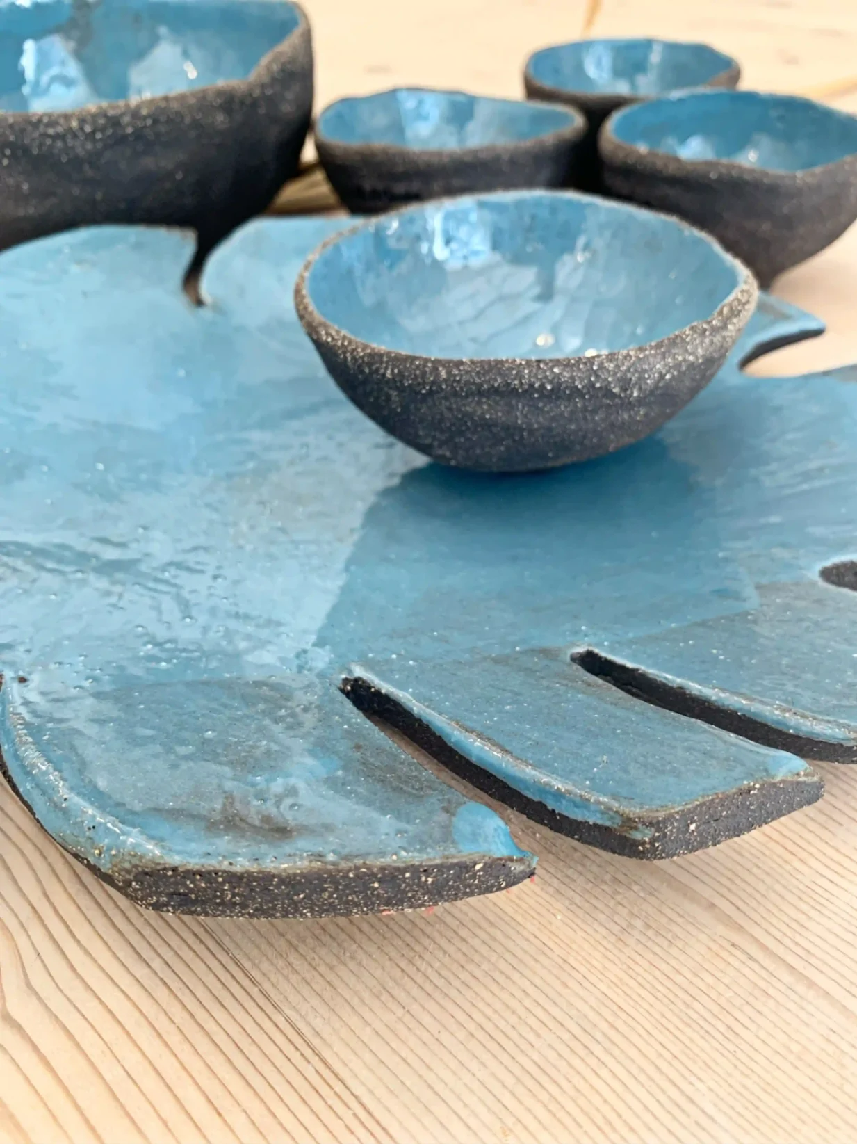 Plateau artisanal en céramique en forme de feuille bleue accompagné de petits bols assortis, finition brute et émaillée.