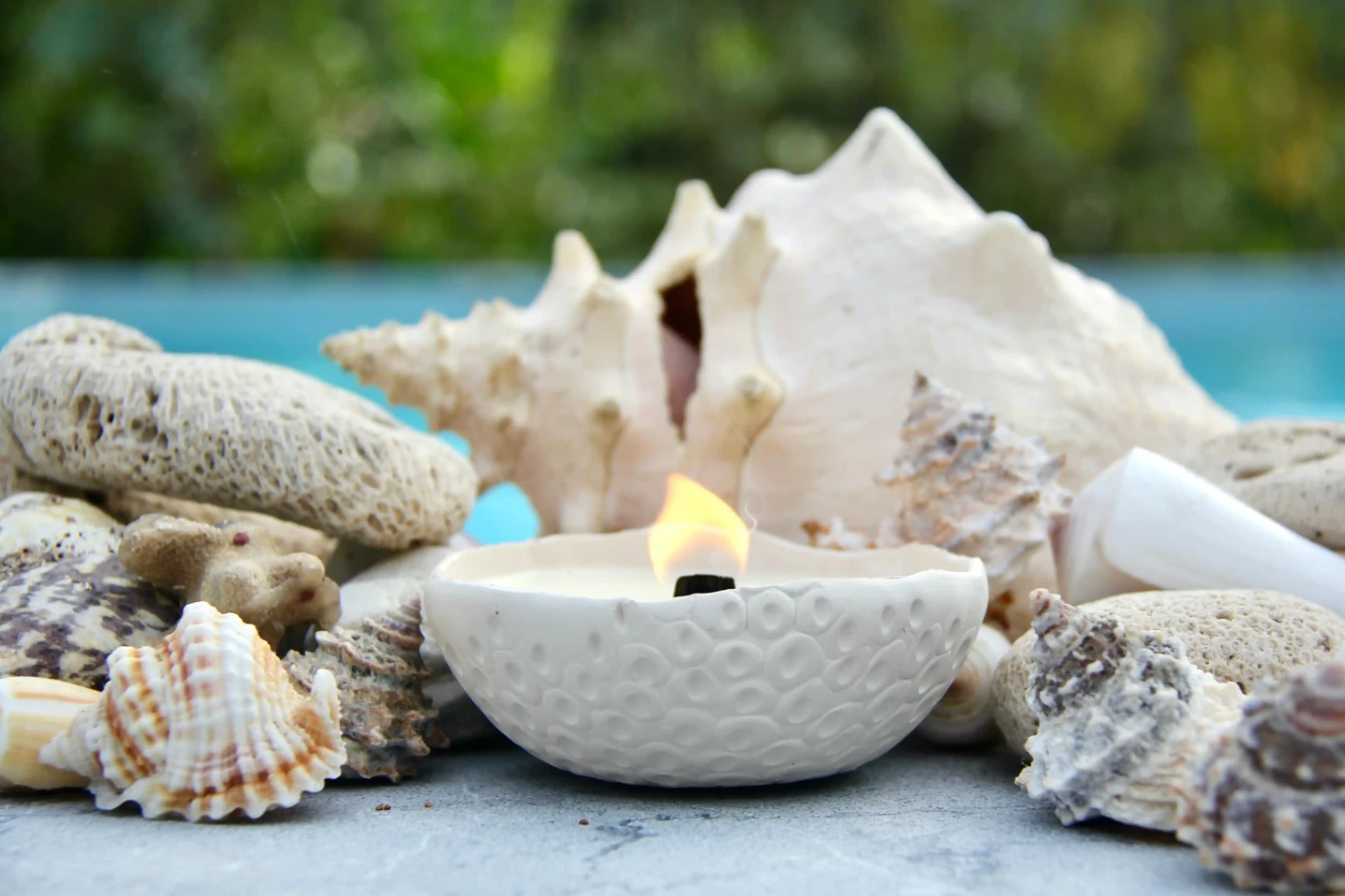 Bougie artisanale en céramique blanche entourée de coquillages et coraux, en extérieur près d’une piscine.