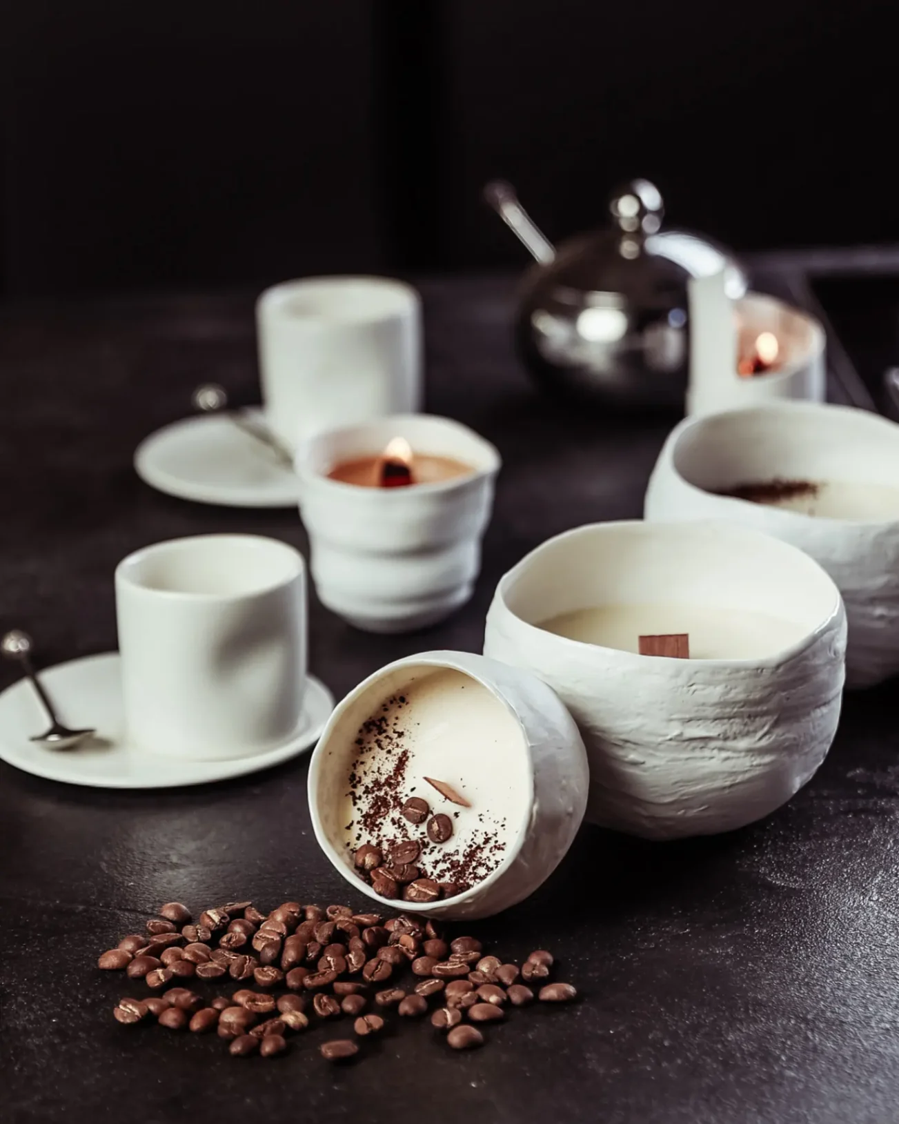 Bougies artisanales en céramique blanche, décor café, entourées de grains de café et de tasses sur table sombre