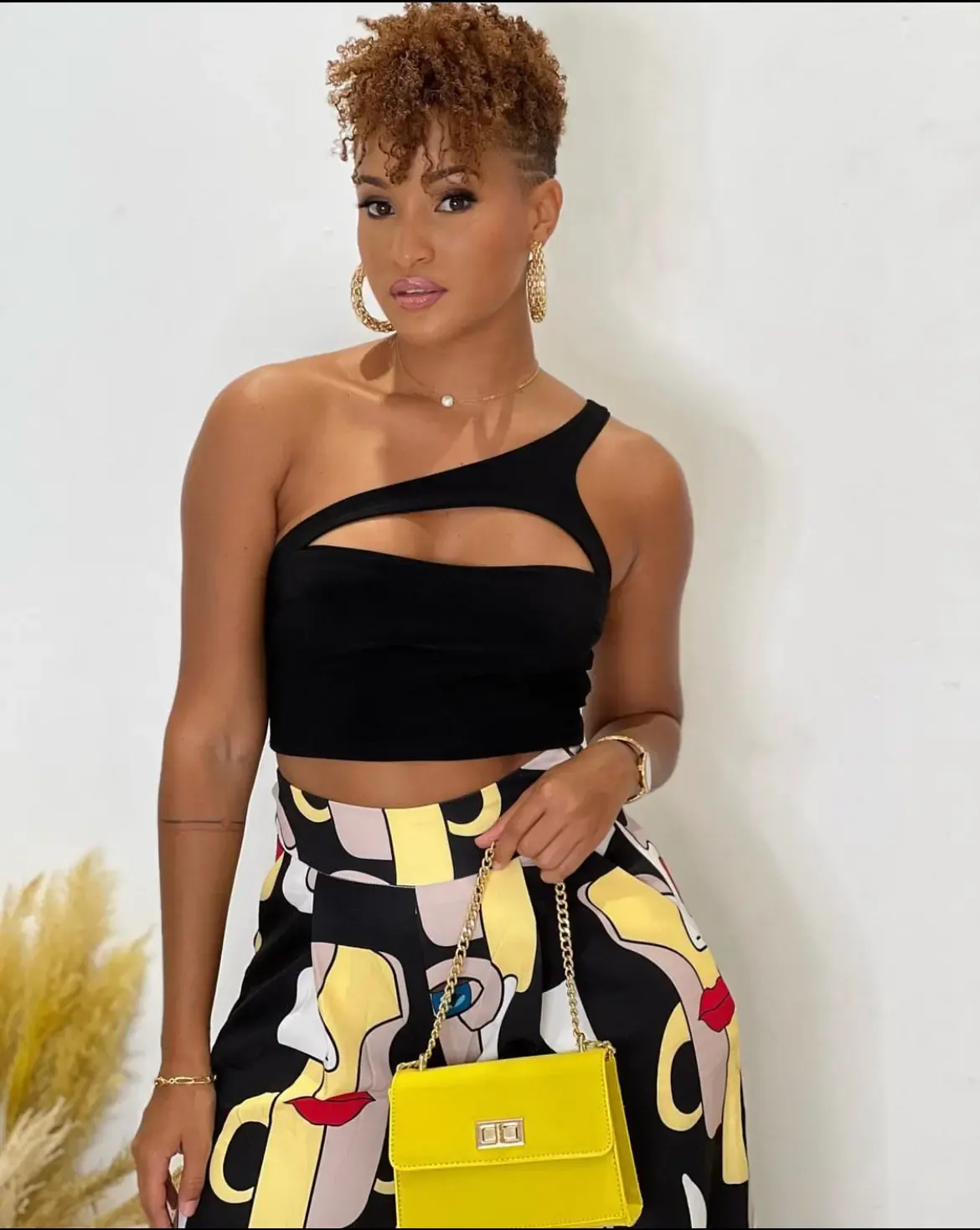 Femme portant un haut noir asymétrique et une jupe imprimée graphique, tenant un petit sac jaune.