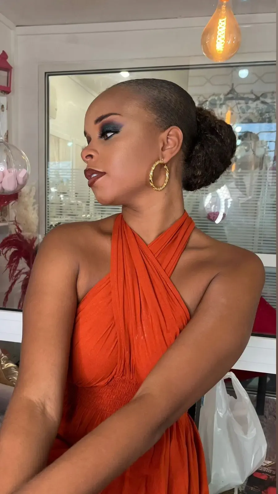 Femme portant une robe orange élégante avec un maquillage glamour et des boucles d’oreilles dorées, posant dans un intérieur lumineux