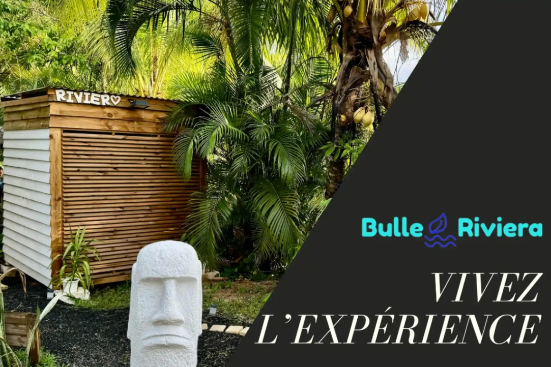 Entrée de Bulle Riviera entourée de végétation tropicale avec sculpture et cabanon en bois, invitant à vivre une expérience immersive