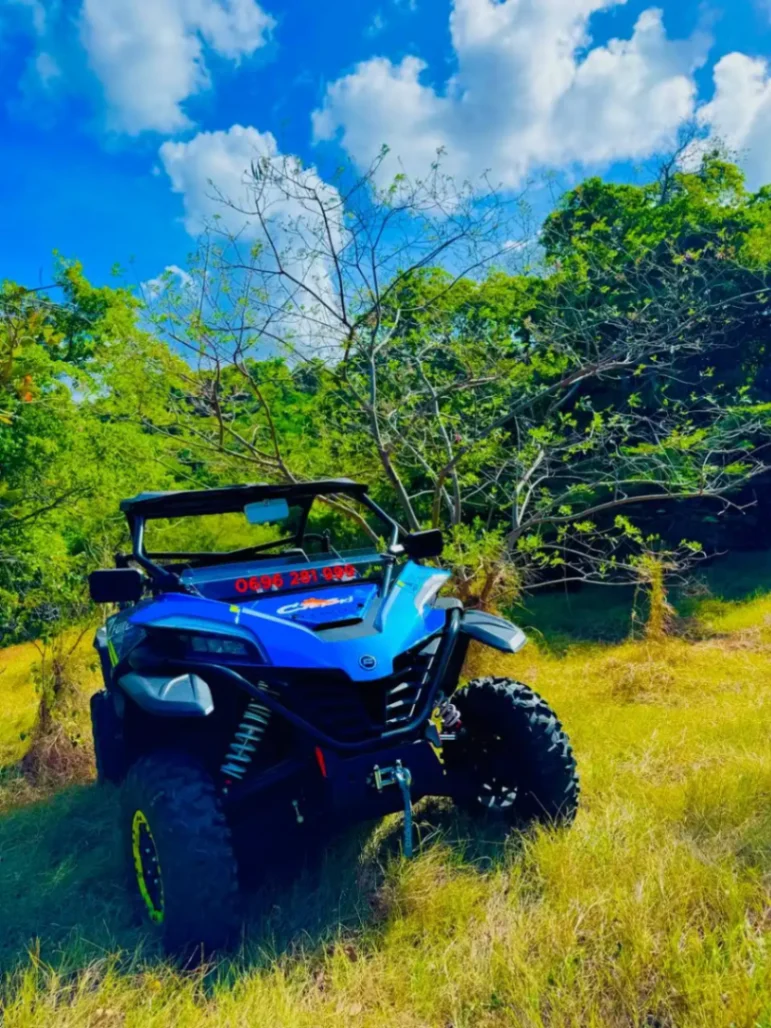 Quad tout-terrain bleu stationné dans une clairière tropicale en Martinique, entouré de végétation luxuriante sous un ciel bleu.