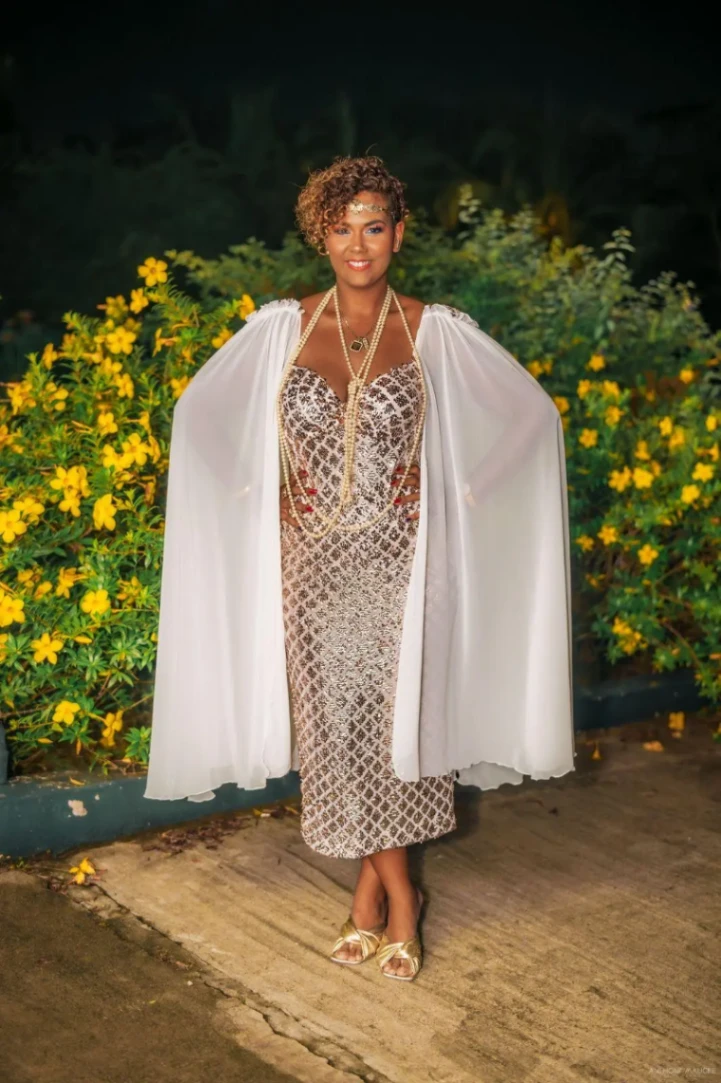 Femme portant une robe élégante claire avec cape blanche, posant devant un jardin aux fleurs jaunes, de nuit.