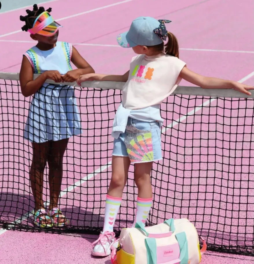 Deux filles discutant sur un terrain de tennis rose, portant des tenues colorées et accessoires estivaux.